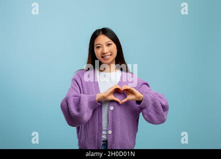 Freundlichkeit und Liebe Konzept. Junge asiatische Dame, die mit Fingern vor ihrer Brust ein Herzzeichen macht, blauer Hintergrund Stockfoto