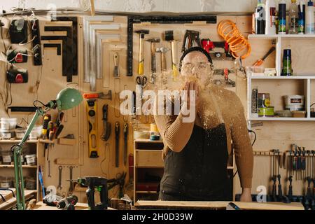 Ein Zimmermann in einer Uhrenmütze, der Staub aus seiner Handfläche in einer großen Werkstatt blies. Anzahl der an der Wand hängenden Werkzeuge. Stockfoto