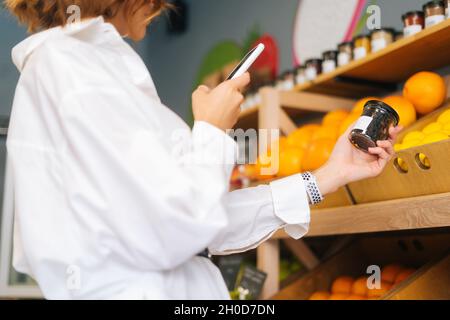 Nahaufnahme einer nicht erkennbaren jungen Frau, die auf dem Markt Produkte auswählt, die über die Theken läuft und mit dem Smartphone Fotos von Gegenständen fotografiert. Stockfoto