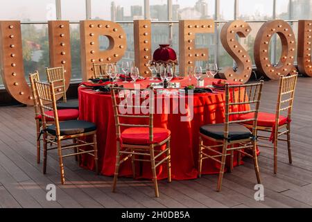 Tische und Stühle für Bankette in Schwarz, Rot und Gold. Eleganter Veranstaltungssaal: Dekor, Tischdecke, Teller, Servietten, Rosen, Brille Stockfoto