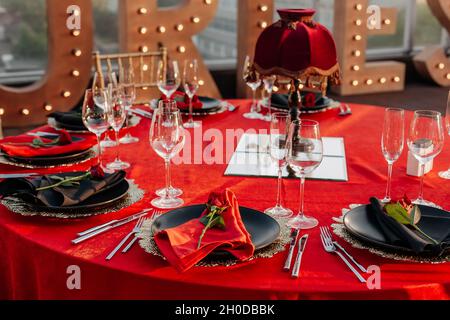 Gäste können an einem Tisch in Schwarz, Rot und Gold Bankett machen. Elegantes Abendessen: Dekor, Tischdecke, Teller mit Servietten und frischen Rosen, Gläser, Cutler Stockfoto