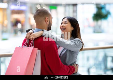 Schönes junges, vielfältiges Paar mit Einkaufstaschen, die sich umarmen und sich im großen Supermarkt angucken Stockfoto