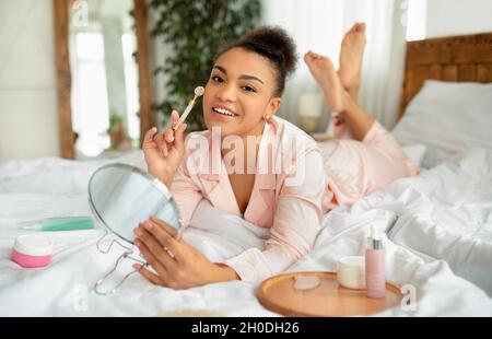 afroamerikanische Dame im Nachtanzug, die mit einem Roller eine Gesichtsmassage macht, auf dem Bett mit Kosmetikprodukten und Spiegel liegt Stockfoto