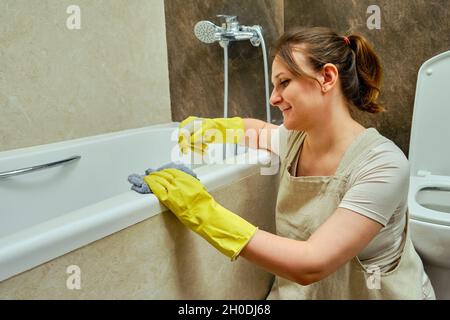 Glückliche Frau putzt das Badezimmer und wäscht die Badewanne. Desinfektion mit gelben Gummihandschuhen im Waschraum Stockfoto