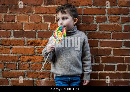 Kleiner Junge mit einem riesigen Lutscher, der sich an einer Ziegelwand lehnt Stockfoto