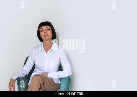 Eine Frau in einem weißen Hemd sitzt auf einem Stuhl vor dem Hintergrund einer grauen Wand. Stockfoto