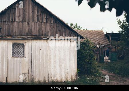 Ein altes rustikales weiß getünchtes Haus mit einem dreieckigen Dach und einem Vorhangfenster, alte Mutter und älterer Sohn im Garten Stockfoto