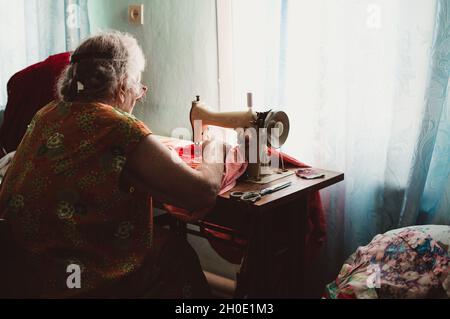 Eine alte, dicke grauhaarige Frau mit lockigem Haar näht an einer Vintage-Retro-Nähmaschine Stockfoto