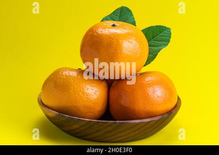 Reife Mandarinenorangen mit grünen Blättern in Holzschüssel isoliert auf gelbem Hintergrund mit Schneideweg. Stockfoto