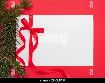 Weihnachtliche Geschenkanhänger weiß blank mit roter Schleife auf rotem Hintergrund. Papiernotiz und Tannengirlande, leerer Raum, Grußkartenvorlage. Neu Ja Stockfoto
