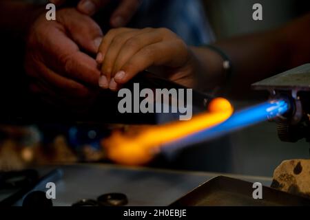 Eine Hand eines Flammenarbeiters und eine Hand eines Kindes während der Herstellung von Glasperlen.Diese Kunst wurde 2020 in die repräsentative Liste des Immateriellen aufgenommen Stockfoto