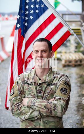 Sgt. Michael Raines, Soldat der Militärpolizei bei der 29. Militärpolizei-Kompanie, Maryland Army National Guard, lächelt am 13. Februar 2021 vor der US-Flagge vor seinem Büro in Camp Bondsteel, Kosovo. Raines wird als Provokant für das Regionalkommando Ost der Kosovo Force eingesetzt. In dieser Position hat Raines die seltene Gelegenheit, MP-Soldaten in einer Mission zur Durchsetzung von Gesetzen und zum Schutz von Truppen in Camp Bondsteel zu führen, anstatt die Kampfunterstützung zu unterstützen, die eine häufiger Mission ist, die den MP-Einheiten der Nationalgarde zugewiesen ist. Er ist verantwortlich für einen Zug aus Verkehrsaufwehrern, Stockfoto