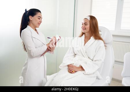 Kosmetologe Beratung weibliche Patientin und Notizen während der medizinischen Termin in der modernen Beauty-Klinik Stockfoto