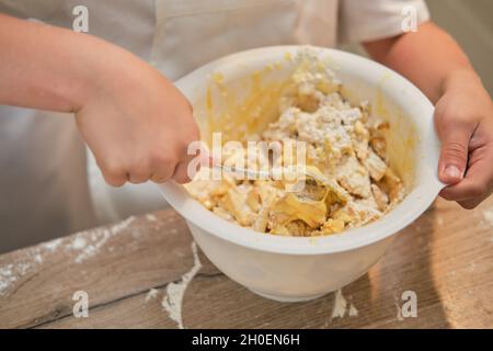 Ein Junge in der Kleidung des Küchenchefs gießt Mehl in den Teig, um Apfelkuchen zu kochen Stockfoto