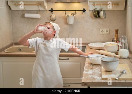 Ein Junge in der Kleidung des Küchenchefs trinkt Milch aus einem Glas, während er in der Küche einen Apfelkuchen kocht Stockfoto