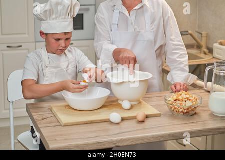 Mutter und Sohn kochen Apfelkuchen in der heimischen Küche. Eine Frau und ein Junge in Küchenhüten und Schürzen kochen mit Gebäck Stockfoto