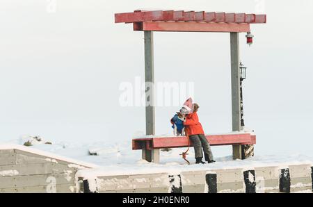 Kid boy trägt Weihnachtsmann Hut wärmt seinen Hund sitzen auf der Bank auf Pier mit gefrorenem weißen Meer im Hintergrund Stockfoto