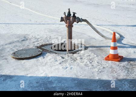 Ein schäbiger Hydrant in einer offenen Luke und ein Verkehrskegel in der Nähe, inmitten einer verschneiten Winterstraße. Stockfoto