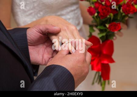 Die Hand eines Mannes legt während der Trauung einen Ring auf seine Braut. Das Mädchen hält einen Strauß roter Rosen in der Hand. Stockfoto