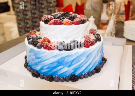 Zweistufige, cremig-weiße und blaue Kuchen steht auf einem Hochzeitstisch. Kuchen dekoriert mit Waldrot und schwarzen Beeren. Stockfoto