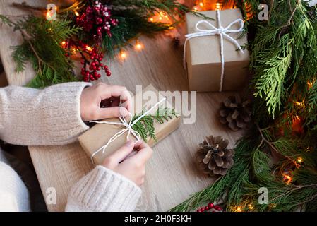 Weibliche Hände wickeln Geschenk in Bastelpapier auf Tisch mit Dekor und Girlande von Lichtern, Weihnachtskonzept, Nahaufnahme. Stockfoto