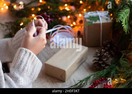 Weihnachtskonzept, weibliche Hände Geschenkverpackung in Bastelpapier auf Tisch mit Dekor und Girlande von Lichtern, Nahaufnahme. Stockfoto