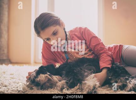 Nettes Mädchen kuscheln Hund auf Teppich im Wohnzimmer neben Fenstern Stockfoto