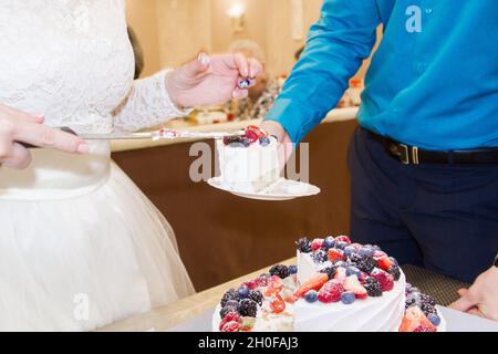 Die Braut und der Bräutigam schneiden auf einer Hochzeitsfeier einen zweistufigen cremeweißen und blauen Kuchen. Kuchen mit Waldbeeren verziert. Stockfoto