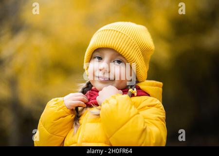 Schönes Outdoor-Herbstporträt von entzückenden 4-jährigen Mädchen, trägt stilvolle gelbe Mütze und Mantel, berühren ihren warmen roten Schal und lächelt an der Kamera Stockfoto