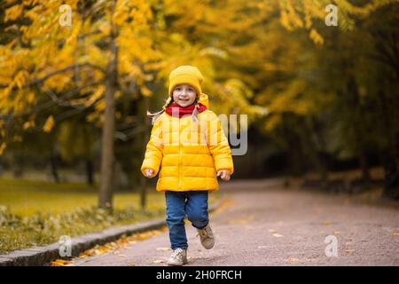 Kleine lustige Mädchen läuft und lächelt auf gelben Bäumen Hintergrund im Herbst Park. Kind auf einem Spaziergang im Herbstpark. Vorschulmädchen läuft und lacht auf der Parkallee Stockfoto