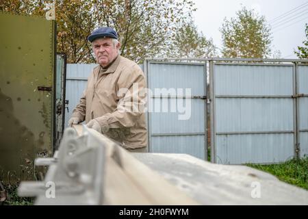 Ein älterer grauhaariger Zimmermann schneidet mit einer Kreissäge ein Brett auf einer Maschine. Selektiver Fokus. Eine rotierende Scheibe der Säge sägt ein Holzbrett entlang. Ein älterer professioneller Mann arbeitet im Freien. Stockfoto