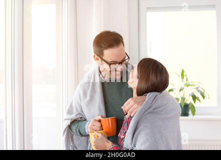 Glückliches junges Paar, das sich unter der Decke in Umarmung neben den Fenstern zu Hause wärmte Stockfoto