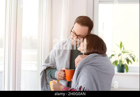 Glückliches junges Paar, das sich unter der Decke in Umarmung neben den Fenstern zu Hause wärmte Stockfoto