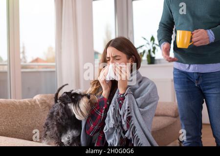 Freund bringt Tee und kümmert sich um Mädchen mit Grippe. Sitzen auf dem Sofa zu Hause bedeckt mit Decke und Nase weht Stockfoto