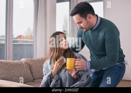 Freund kümmert sich um Mädchen mit Grippe. Sitzen auf dem Sofa zu Hause mit Decke bedeckt und Tee trinken Stockfoto