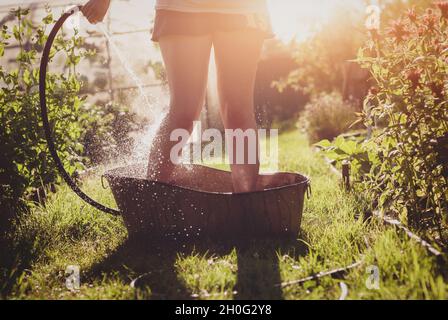 Baden im Sommergarten - die Frau gießt kaltes Wasser auf ihre Füße aus dem Gartenschlauch, der in einem eisernen Bad auf dem Gartenweg im Sonnenlicht steht Stockfoto