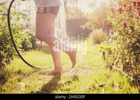 Frau gießt Wasser auf ihre Füße aus Gartenschlauch im Hinterhof, Spaß an einem heißen Sommertag Stockfoto