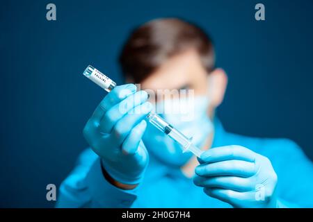Ein Arzt in einer medizinischen Maske und Handschuhen, in einer blauen Uniform gekleidet, wählt den Impfstoff in eine Spritze Stockfoto