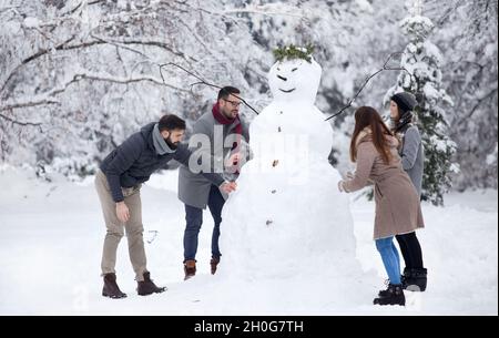 Zwei junge Paare, die sich während der Winterferien beim Bau eines großen Schneemanns im Park amüsieren Stockfoto