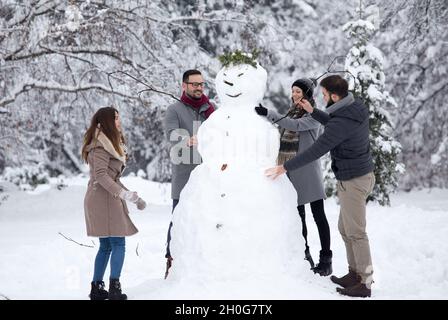 Zwei junge Paare, die sich während der Winterferien beim Bau eines großen Schneemanns im Park amüsieren Stockfoto