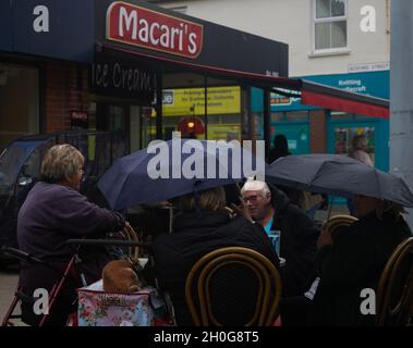 Die Leute genießen ihre Zeit zusammen und trinken draußen auf der Terrasse des Macaris Cafés in Bognor Regis, die vor dem Regen mit Sonnenschirmen geschützt sind. Stockfoto