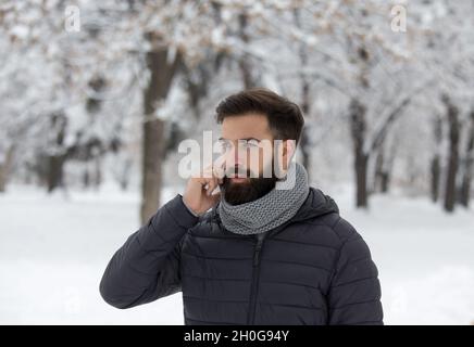 Hübscher junger Mann mit Bart, der im Wald mit Schnee auf dem Handy spricht Stockfoto