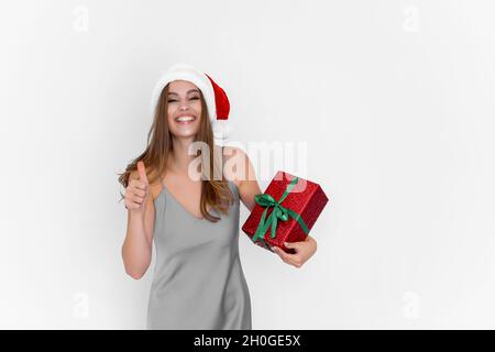 Glücklich lächelnd aufgeregt Santa Mädchen hält Geschenkbox, während Gesten Daumen nach oben tun ausgezeichnet. Feiern Sie. Stockfoto