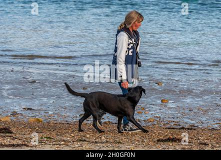 Dame oder Hündin mittleren Alters, die einen Hund am Strand entlang läuft. Hundeausbildung am Meer, Hundespaziergang am Strand, Spaziergang mit einem Hund am Strand. Stockfoto