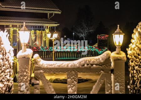Weihnachtliche Lichtdekoration auf Villa und Holzzaun, bunte Lichter leuchten. Verschwommenes Gartentor mit zwei weißen Laternen im Vordergrund Stockfoto