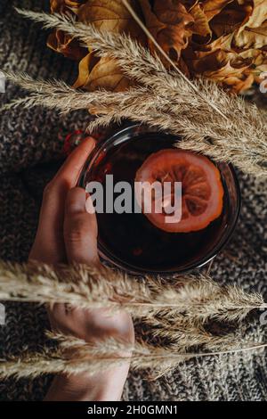 Weibliche Hand hält eine Tasse heißen Tee mit Zitrone und trockener Pflanze auf einem warmen Strickstoff. Herbst gemütliches Zuhause Stillleben. Vertikale Aufnahme Stockfoto