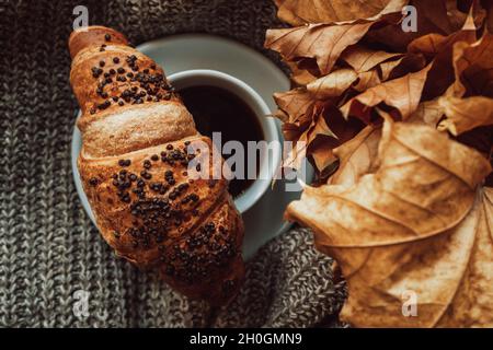 Eine Tasse schwarzen Morgenkaffee mit einem Schokoladencroissant in einem gemütlichen herbstlichen Stillleben. Leckeres Essen. Ästhetik im Detail Stockfoto