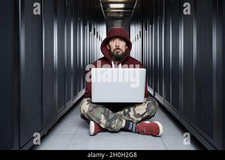 Der Hacker in einer Haube sitzt mit dem Computer unter den Servern, der Einbrecher. Speicherung von Daten, cloudy Services, Systembruch, Angriff von Hackern Stockfoto