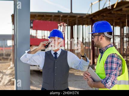 Wütender leitender Ingenieur mit Helm, der am Telefon telefoniert und neben einem jungen Arbeiter auf der Baustelle steht Stockfoto