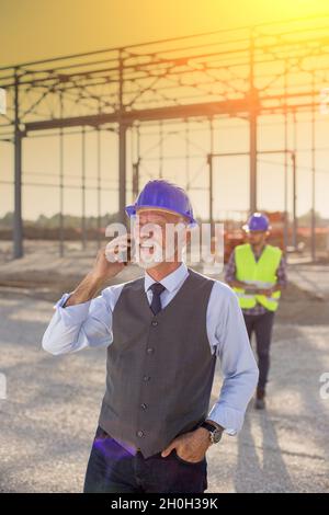 Portrait eines Vorarbeiters mit Helm, der auf dem Handy vor der Metallkonstruktion auf der Baustelle spricht Stockfoto